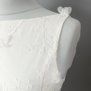 May include: White sleeveless dress with delicate floral embroidery and tulle flower accents on the shoulders.