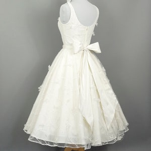 May include: A white wedding dress with a fitted bodice and a full skirt. The dress has a satin bow at the waist and delicate floral embroidery throughout.