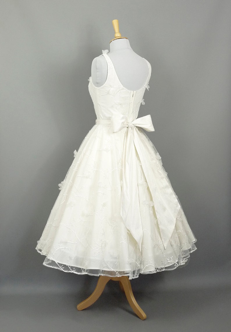 May include: A white wedding dress with a fitted bodice and a full skirt. The dress has a satin bow at the waist and delicate floral embroidery throughout.