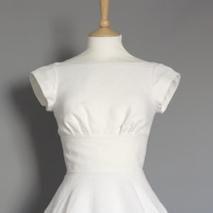 Puede incluir: Un vestido blanco con un corpi&ntilde;o ajustado y una falda completa. El vestido tiene un escote redondo y mangas cortas.