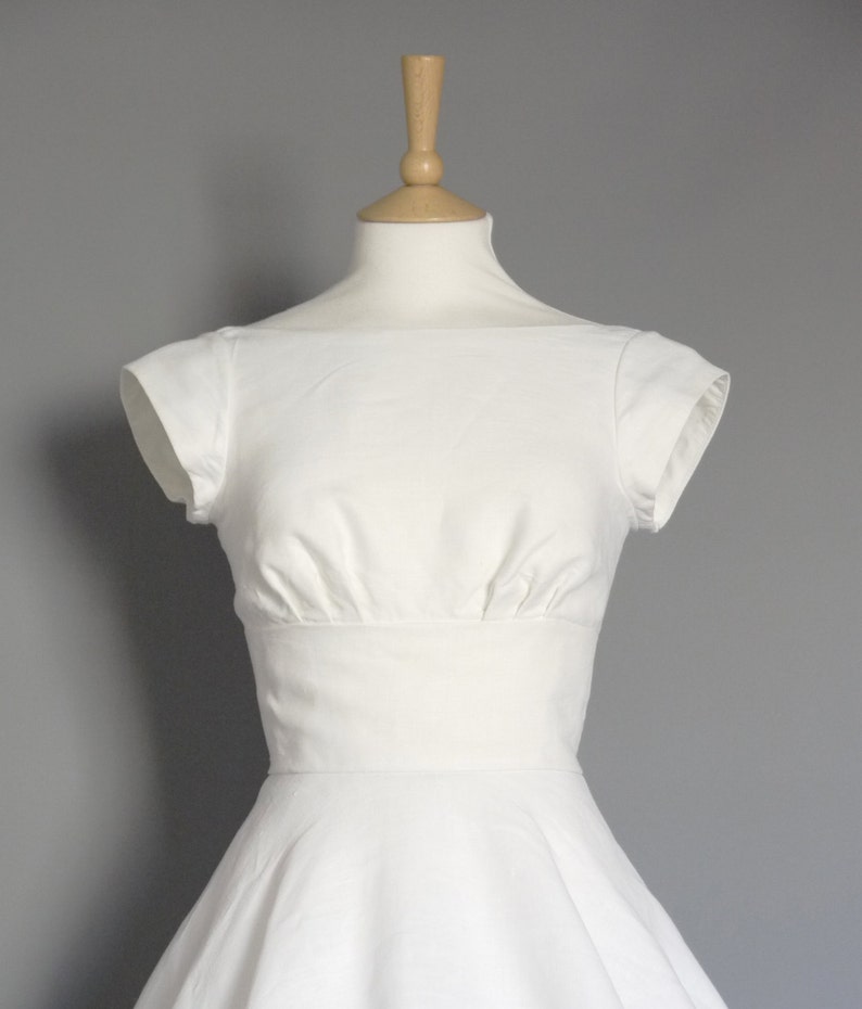 Puede incluir: Un vestido blanco con un corpi&ntilde;o ajustado y una falda completa. El vestido tiene un escote redondo y mangas cortas.