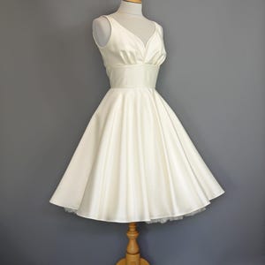 Puede incluir: Vestido de satén color marfil con corpiño con cuello en V y falda acampanada. El vestido se exhibe en un maniquí de madera. La falda tiene un delicado ribete de encaje en el dobladillo. El vestido es de estilo vintage.