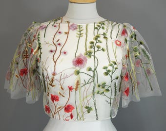 May include: A white, floral embroidered top with sheer, short sleeves. The embroidery features red, pink, and green flowers and foliage. The top is displayed on a mannequin against a gray background.