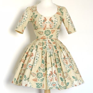 Op de afbeelding: Een vintage, crèmekleurige jurk met een bloemenprint met groene bladeren, oranje vogels en roze bloemen. De jurk heeft een getailleerd bovenlijf en een volle rok met plooien.