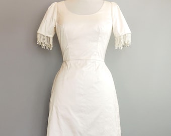 May include: Ivory-colored vintage dress with a scoop neckline and short sleeves. The sleeves are adorned with beaded fringe. The dress has a fitted bodice and a slightly flared skirt, displayed on a mannequin against a gray backdrop.