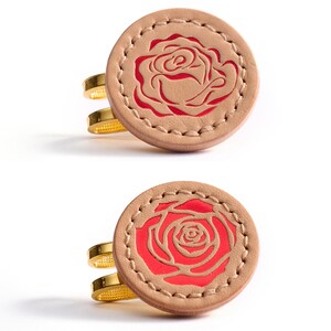 May include: Two rings with a circular design featuring a red rose on a tan leather background. The rings have a gold-colored band. The rose design is stitched into the leather, creating a detailed and elegant look.