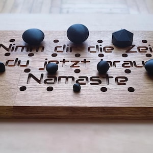 May include: Wooden board with engraved text and various black shapes. The text reads "Nimm dir die Zeit die du jetzt brauchst Namaste". The board has several small, round holes and is made of light brown wood.