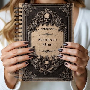 Peut inclure: Un carnet à spirales au design vintage. La couverture présente un crâne et des ornements floraux, avec les mots "Memento Mori" au centre. Le carnet a une palette de couleurs marron foncé et beige, avec une esthétique gothique.
