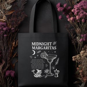 Puede incluir: Bolso tote negro con las palabras "MIDNIGHT MARGARITAS" en escritura blanca. El diseño incluye una copa de cóctel, una luna creciente, una rana y ilustraciones botánicas. El bolso tiene un asa negra.