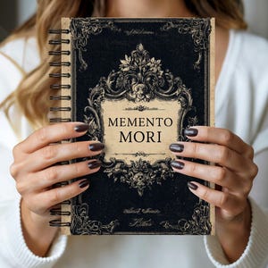 Op de afbeelding: Een spiraalgebonden notitieboek met een zwarte kaft met sierlijke, vintage-achtige krullen en de woorden "MEMENTO MORI" in een schreefletter. Het notitieboek heeft een crèmekleurig label en een donkerbruine spiraalbinding.