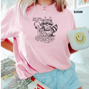 May include: A light pink t-shirt with a black and white illustration of an angry raccoon pouring ice cubes from a container. The text "STAY FERAL ABOLISH ICE" is above the raccoon. A white mug with a yellow smiley face is in the image.