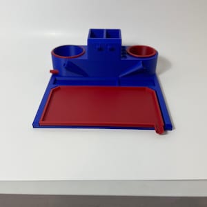 May include: A two-tone organizer in blue and red. The organizer has a rectangular base with a red tray. Above the tray are two circular holders, a small rectangular compartment, and two angled chutes.