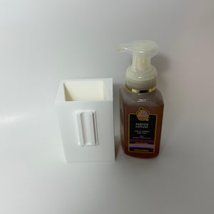 May include: A white rectangular soap dispenser holder with a rectangular design on the front, next to a bottle of Pumpkin Cupcake scented foaming hand soap. The soap bottle is brown with a gold pump.