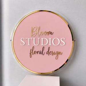 May include: A circular sign featuring a light pink background with a gold metallic border. The text Bloom Studios is displayed in gold script and white block letters, with the words floral design in gold script below.