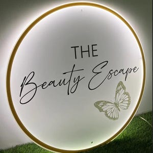 May include: A round, illuminated sign with a gold border. The sign features the words "THE Beauty Escape" in elegant script, along with a butterfly graphic. The background is white, and the sign is lit with a soft glow.