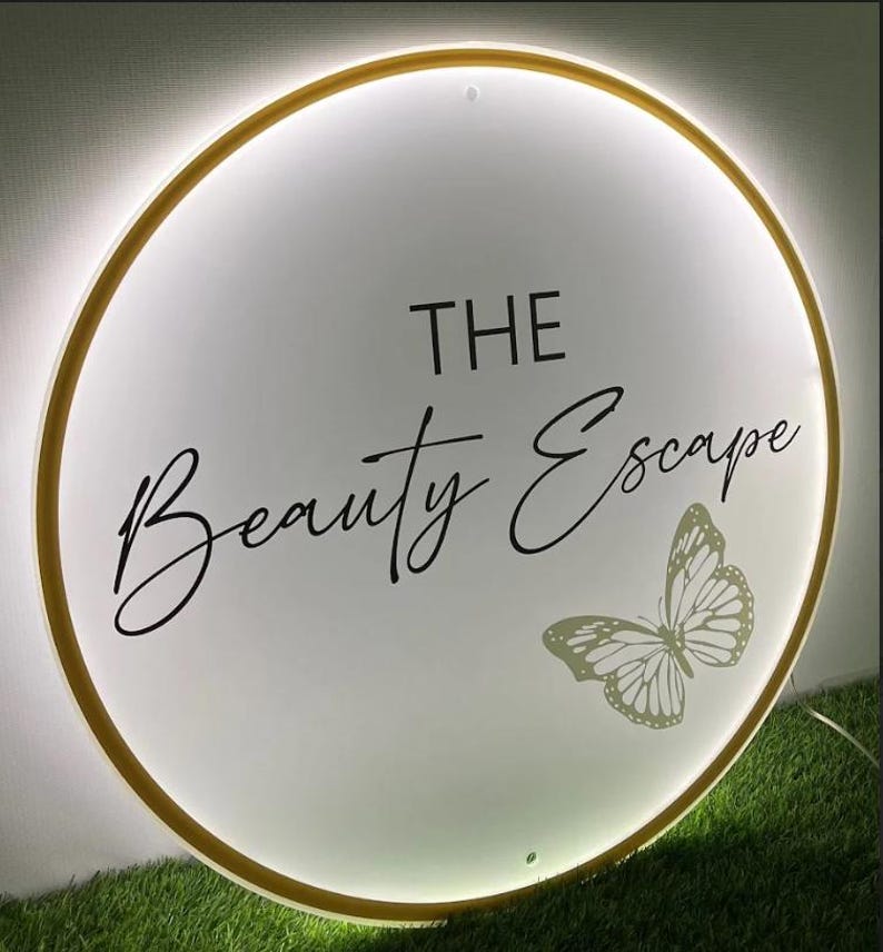 May include: A round, illuminated sign with a gold border. The sign features the words "THE Beauty Escape" in elegant script, along with a butterfly graphic. The background is white, and the sign is lit with a soft glow.