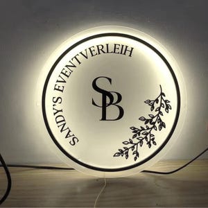 May include: A round, illuminated sign with the text "SANDY'S EVENT VERLEIH" in a circular pattern. The center features the intertwined letters "SB" and a decorative leaf design. The sign has a black border and is lit from within.