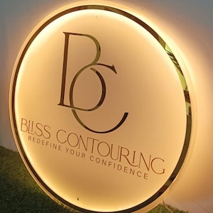 May include: A round, illuminated sign with a gold trim and a cream-colored face. The sign features the letters "BC" in a stylized font, with the words "BLISS CONTOURING" and "REDEFINE YOUR CONFIDENCE" in a serif font.