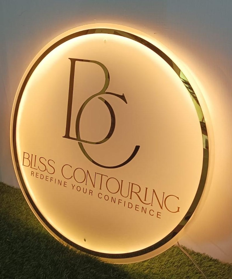 May include: A round, illuminated sign with a gold trim and a cream-colored face. The sign features the letters "BC" in a stylized font, with the words "BLISS CONTOURING" and "REDEFINE YOUR CONFIDENCE" in a serif font.