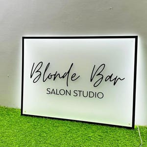 May include: A rectangular sign with a black frame and a white illuminated surface. The sign reads "Blonde Bar SALON STUDIO" in elegant black script. The sign is placed on a bed of green artificial grass.