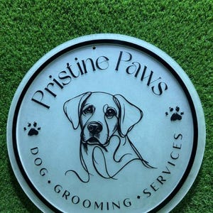 May include: A circular sign with a frosted finish featuring a black line drawing of a dog's face in the center. The text "Pristine Paws" is arched above, with "DOG GROOMING SERVICES" below, accompanied by small black paw print icons.