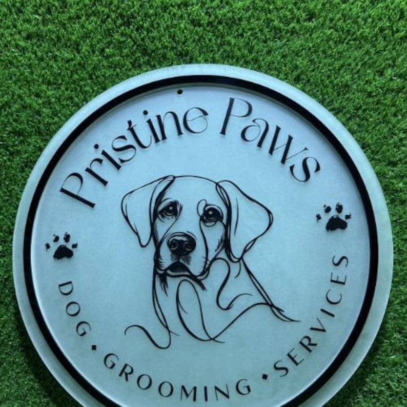 May include: A circular sign with a frosted finish featuring a black line drawing of a dog's face in the center. The text "Pristine Paws" is arched above, with "DOG GROOMING SERVICES" below, accompanied by small black paw print icons.