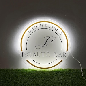 May include: A circular, illuminated acrylic sign featuring the text JAYDAH & JANIYA at the top, a large J2 monogram in the center, and the words BEAUT&Eacute; BAR at the bottom. The sign has a gold-colored circular border and emits a bright white glow.