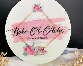 May include: A circular sign with the words "Bake-A-Holic" in a cursive font, and "BY MADIHA GAGAI" underneath. The design includes a pink watercolour wash, floral elements, and a teacup illustration, all on a white background.