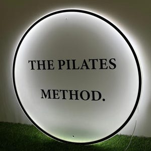 May include: A round, illuminated sign with the text "THE PILATES METHOD." The sign has a black border and a bright white interior. The sign is mounted on a wall above a patch of green grass.