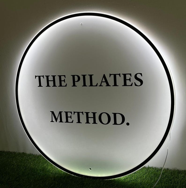 May include: A round, illuminated sign with the text "THE PILATES METHOD." The sign has a black border and a bright white interior. The sign is mounted on a wall above a patch of green grass.