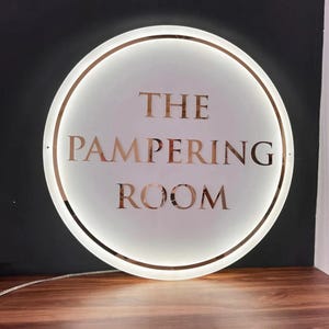 May include: A round, illuminated sign with the text "THE PAMPERING ROOM" in a rose gold color. The sign has a white center with a rose gold border and a warm, inviting glow, perfect for a spa or salon.