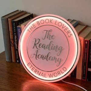 May include: A round, illuminated sign with the text "The Book Lovers" and "The Reading Academy Eternal World" in a soft pink glow. The sign is next to a stack of books, creating a cozy, literary ambiance.