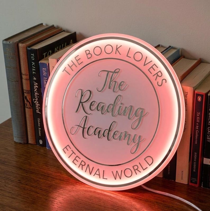 May include: A round, illuminated sign with the text "The Book Lovers" and "The Reading Academy Eternal World" in a soft pink glow. The sign is next to a stack of books, creating a cozy, literary ambiance.