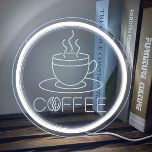 May include: A round, illuminated sign with a white outline and a coffee theme. The sign features a coffee cup with steam and the word "COFFEE" in a stylized font. The sign is lit with a bright white light.