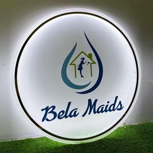 May include: Circular illuminated sign with the text "Bela Maids" in a cursive blue font. The sign features a logo of a house inside a water droplet, with a silhouette of a person cleaning. The sign has a white background and a gold trim.