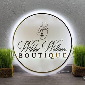 May include: A round, illuminated sign with the text "Wilder Wellness BOUTIQUE" in a stylish font. The sign features a minimalist line drawing of a face and a gold border. Two small green plants in brown pots flank the sign.