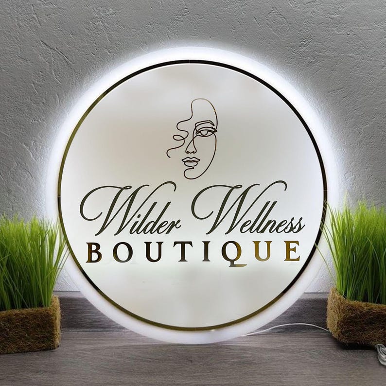 May include: A round, illuminated sign with the text "Wilder Wellness BOUTIQUE" in a stylish font. The sign features a minimalist line drawing of a face and a gold border. Two small green plants in brown pots flank the sign.