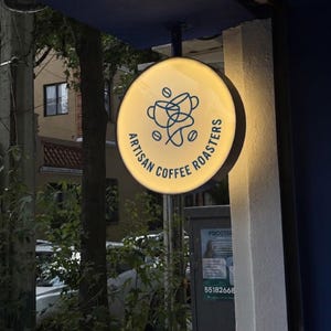 May include: A round, illuminated sign with the words "ARTISAN COFFEE ROASTERS" in a circular design. The sign has a yellow background with a blue coffee cup and bean graphic. The sign is mounted on a blue structure.
