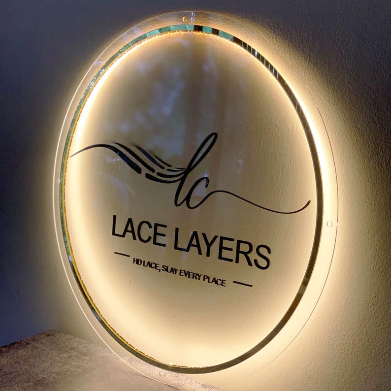 May include: A round, illuminated sign with the text "LACE LAYERS" in black script. The sign has a clear, circular face with a glowing, warm-toned light around the edge. Below the main text is the phrase "HD LACE, SLAY EVERY PLACE".