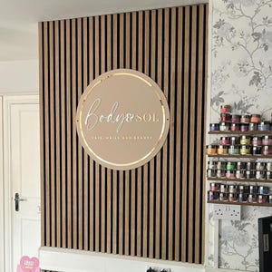 May include: A circular beige sign with gold lettering reading Body & SOL HAIR, NAILS AND BEAUTY mounted on a wall of vertical wood slats. The sign features a thin gold circular border and is positioned centrally on the slatted wooden paneling.