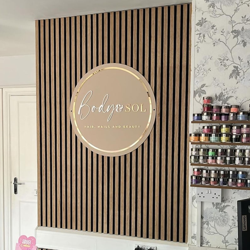 May include: A circular beige sign with gold lettering reading Body & SOL HAIR, NAILS AND BEAUTY mounted on a wall of vertical wood slats. The sign features a thin gold circular border and is positioned centrally on the slatted wooden paneling.