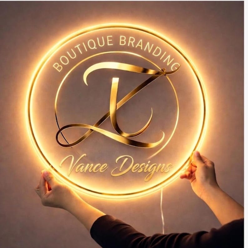 May include: A round, illuminated sign with the text "BOUTIQUE BRANDING" and "Vance Designs" in gold lettering. The sign features a gold-colored "Z" design in the center and is held by two hands. The sign has a warm, yellow glow.