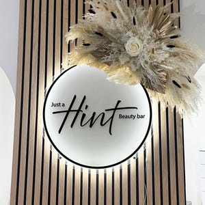 Könnte beinhalten: Ein rundes, beleuchtetes Schild mit dem Schriftzug "Just a Hint Beauty bar" in schwarzer Schrift. Das Schild befindet sich vor einem Hintergrund aus vertikalen Holzlatten. Über dem Schild befindet sich eine dekorative Anordnung aus getrockneten Blumen und Gräsern in neutralen Tönen.