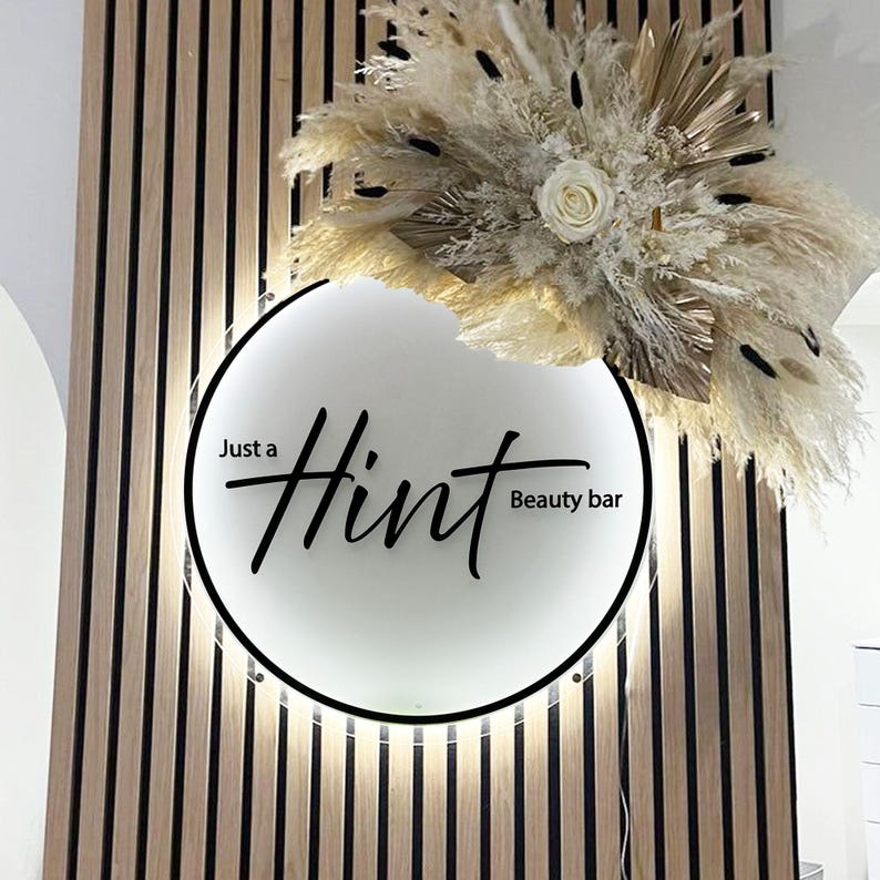 May include: A round, illuminated sign with the words "Just a Hint Beauty bar" in black script. The sign is set against a backdrop of vertical wooden slats. Above the sign is a decorative arrangement of dried flowers and grasses in neutral tones.