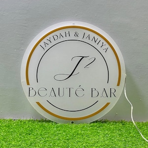 May include: A circular white sign with a gold border featuring the black text JAYDAH & JANIYA, a large stylized J2 monogram in the center, and the words BEAUT&Eacute; BAR at the bottom. A thin white power cord extends from the right side.