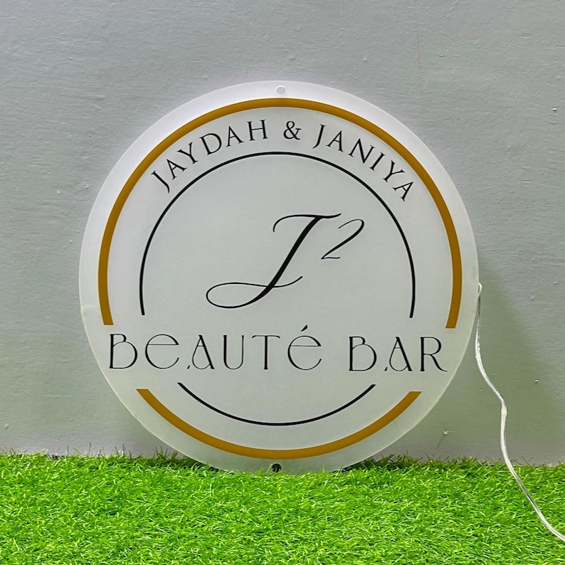 May include: A circular white sign with a gold border featuring the black text JAYDAH & JANIYA, a large stylized J2 monogram in the center, and the words BEAUT&Eacute; BAR at the bottom. A thin white power cord extends from the right side.