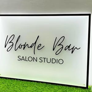 May include: A rectangular sign with a black frame and the words "Blonde Bar SALON STUDIO" in elegant black script. The sign is illuminated with a soft glow and set against a backdrop of green artificial grass.