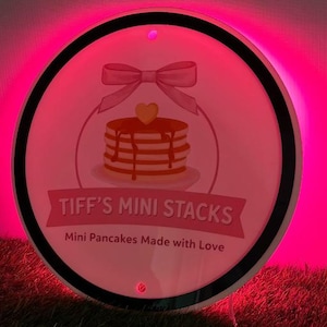 May include: A round sign with a pink background and a black border. The sign features a stack of pancakes with syrup, a bow, and the text "TIFF'S MINI STACKS" and "Mini Pancakes Made with Love".