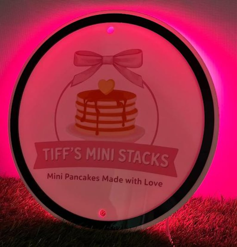 May include: A round sign with a pink background and a black border. The sign features a stack of pancakes with syrup, a bow, and the text "TIFF'S MINI STACKS" and "Mini Pancakes Made with Love".