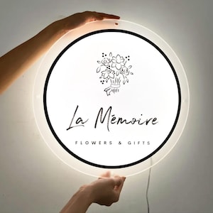 May include: A round, illuminated sign with a white face and black border. The sign features the text "La M&eacute;moire FLOWERS & GIFTS" and a floral illustration. The sign is held by two hands.